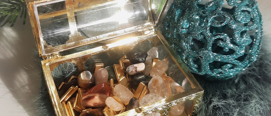 Eine goldene Truhe, gefüllt mit verschiedenen Kristallen und Edelsteinen, liegt auf einem Pelztuch. Daneben befindet sich eine türkisfarbene Dekorationskugel. Hinter der Truhe steht eine Kerze in einem Glashalter und einige Tannenzweige.