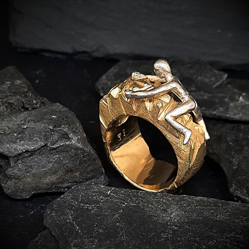 Ein goldener Ring mit einer silbernen Klettererfigur, die auf einer Felsenoberfläche ruht. Der Ring hat eine strukturierte Oberfläche mit schwarzem Hintergrund.