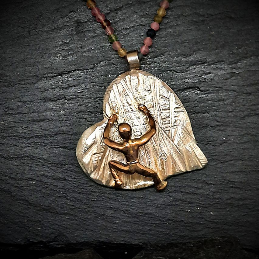 Ein goldener Herzanhänger mit einer bronzefarbenen Figur, die Yoga macht, auf einer dunklen Oberfläche.