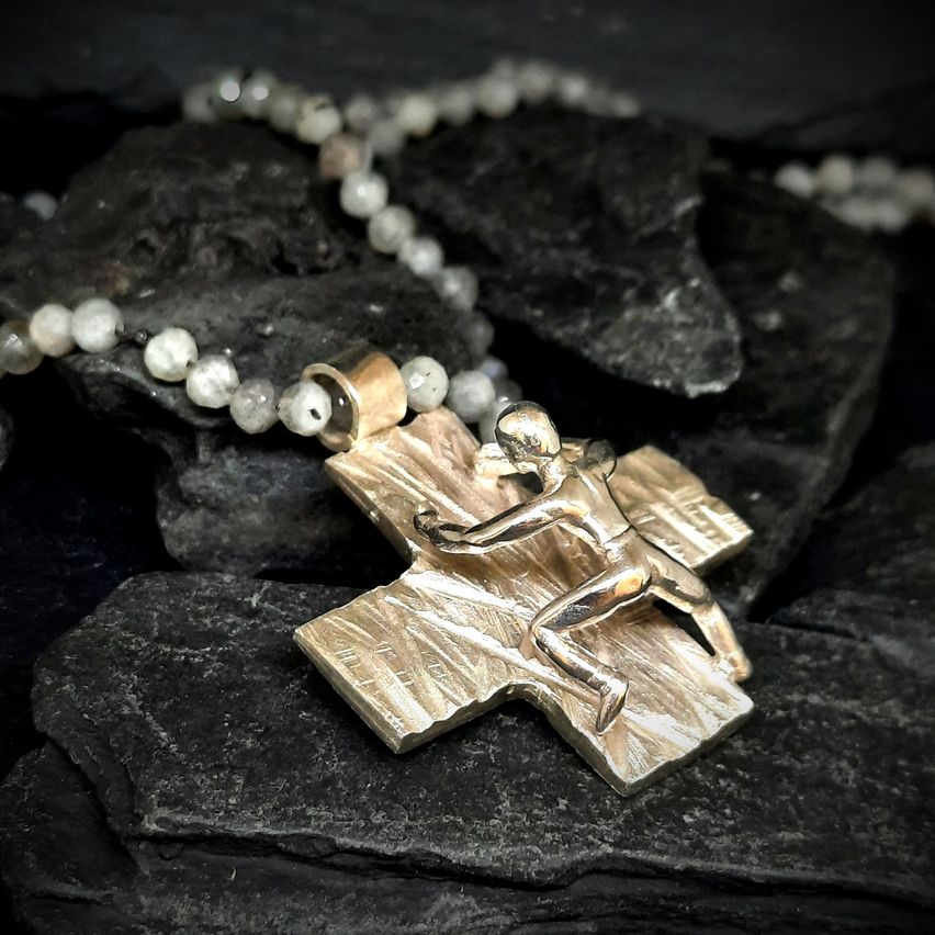 Eine silberne Kreuz-Anhänger mit einer laufenden Figur, ruht auf einer Perlenkette aus weißen Perlen und dunklen Steinen.