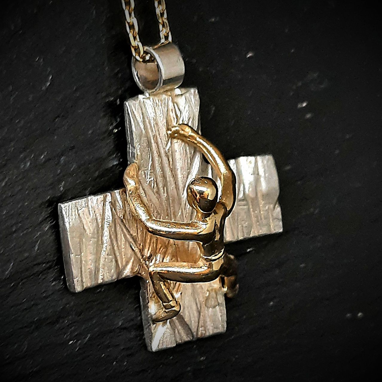 Eine goldene Figur klettert auf einen strukturierten silbernen Kreuzanhänger, befestigt an einer Kette, vor einem dunklen Hintergrund.