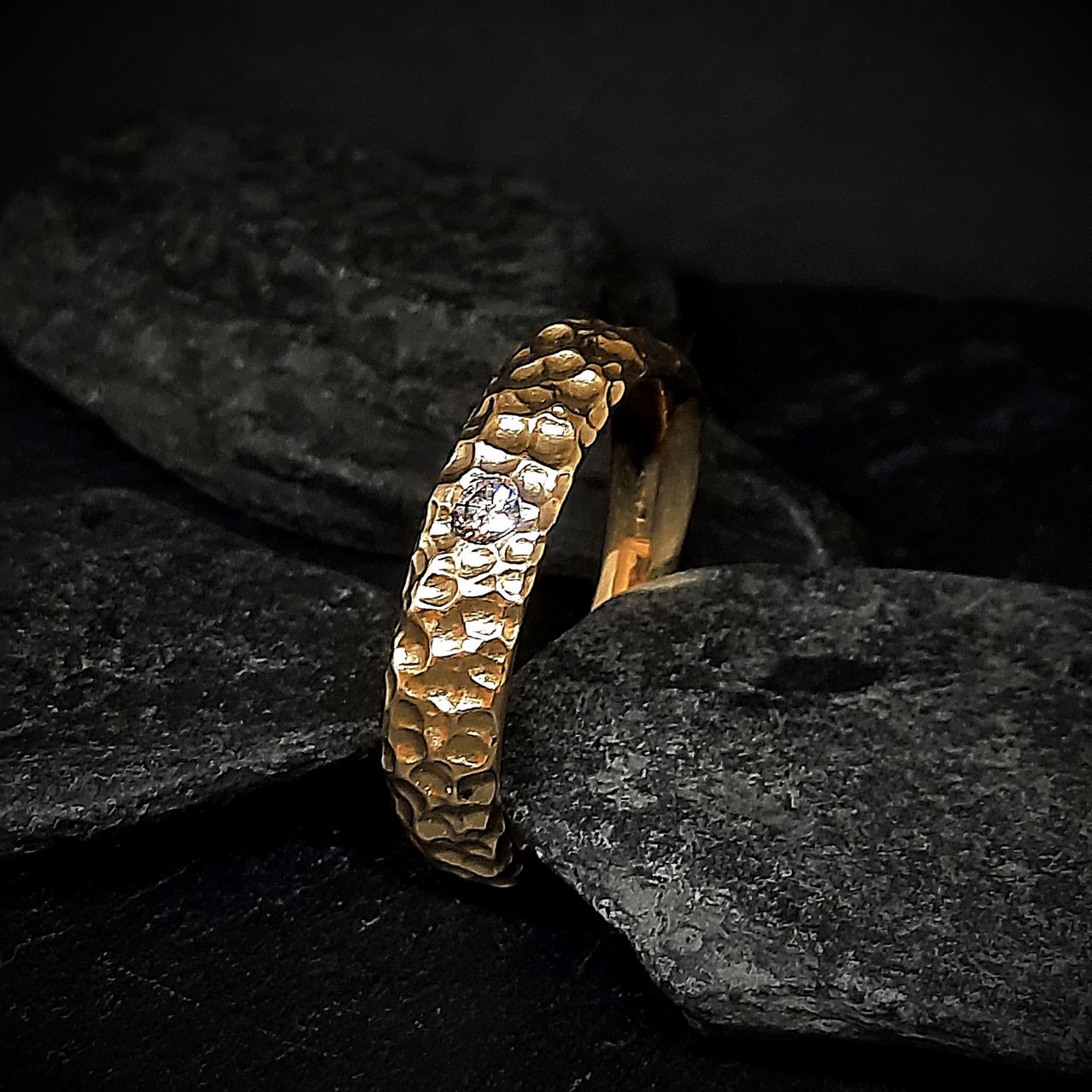 Ein goldener Ring mit Textur und einem einzelnen Diamanten ruht auf dunklen Steinen. Die Textur des Rings kontrastiert mit den glatten Steinen.