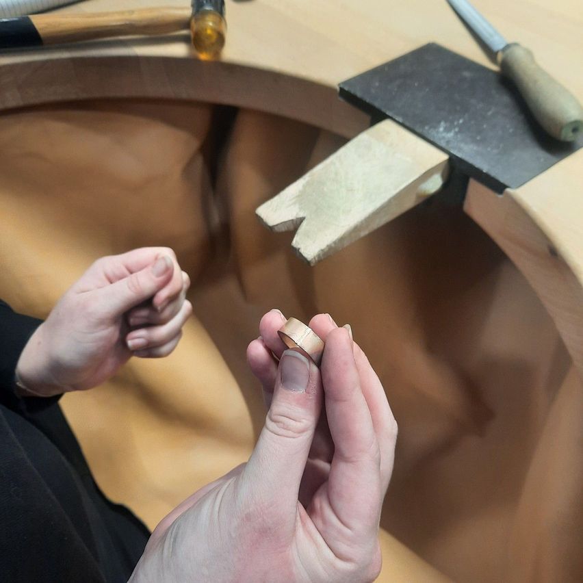 Eine Person fertigt einen Ring in einer Werkstatt, mit einem hölzernen Arbeitstisch, einem Hammer und einem Meißel. Die Person hält ein kleines Metallstück und untersucht es genau.