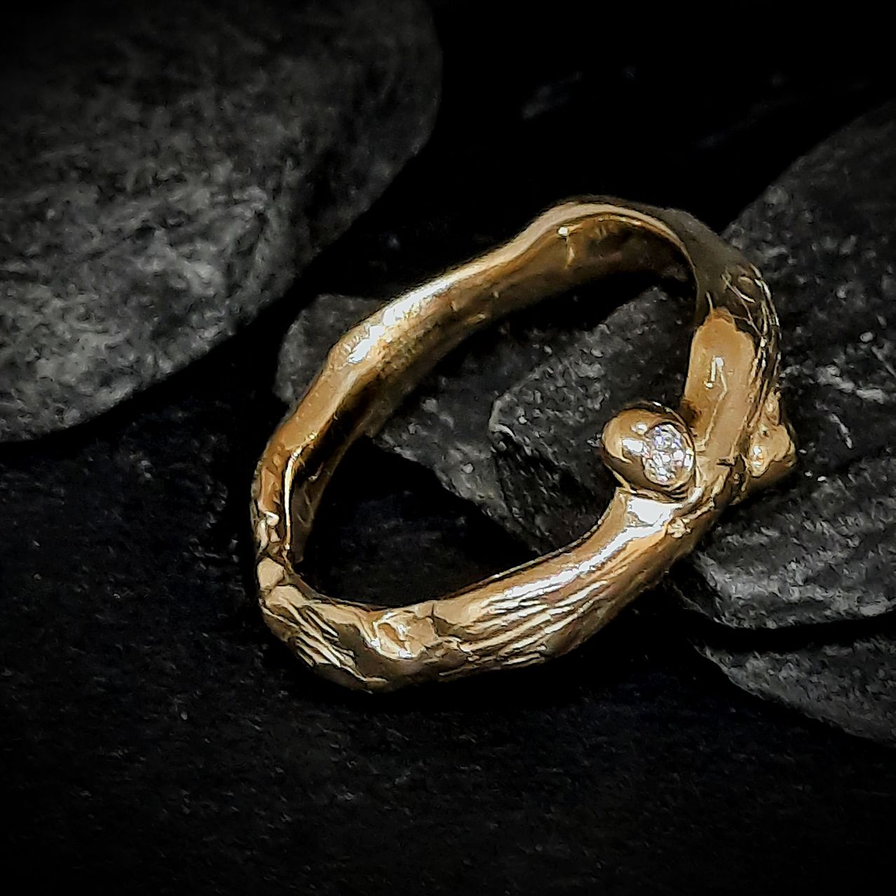 A golden ring with a tree branch design and a diamond sits on black rocks.