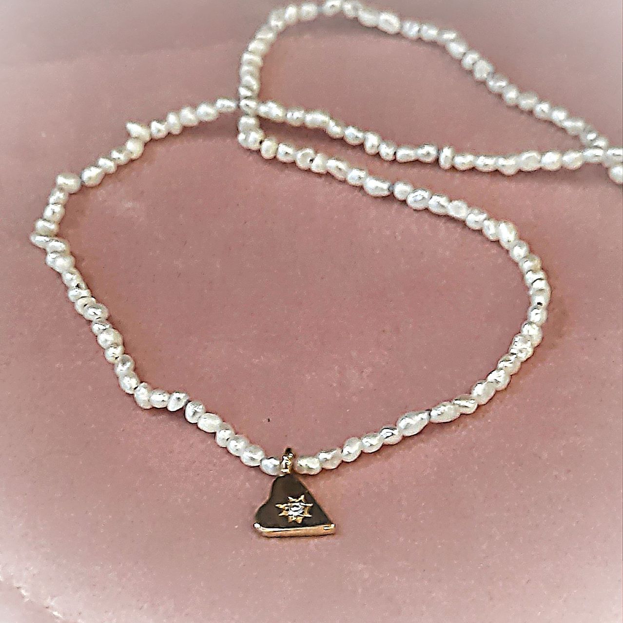 A white pearl necklace with a golden pendant featuring a sun and diamond is displayed on a pink surface.