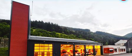 A fire station building with red exterior, multiple fire trucks parked inside, set against a backdrop of hills and clear sky, with the date August 8, 2026, written in red.