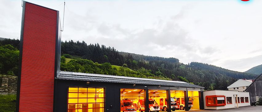 A fire station building with red exterior, multiple fire trucks parked inside, set against a backdrop of hills and clear sky, with the date August 8, 2026, written in red.