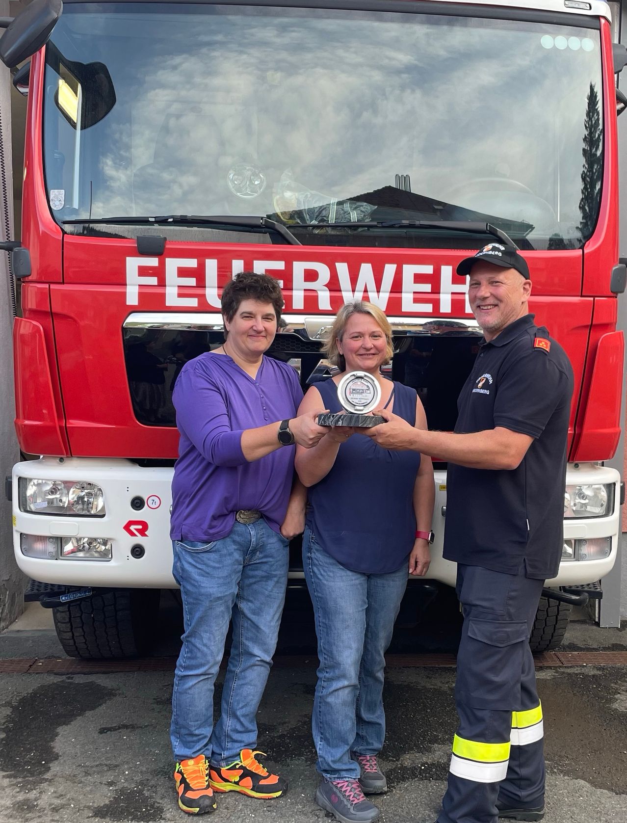 Drei Personen posieren mit einem Preis vor einem roten Feuerwehrauto. Das Auto steht 'Feuerwehr' und hat ein Logo an der Seite.