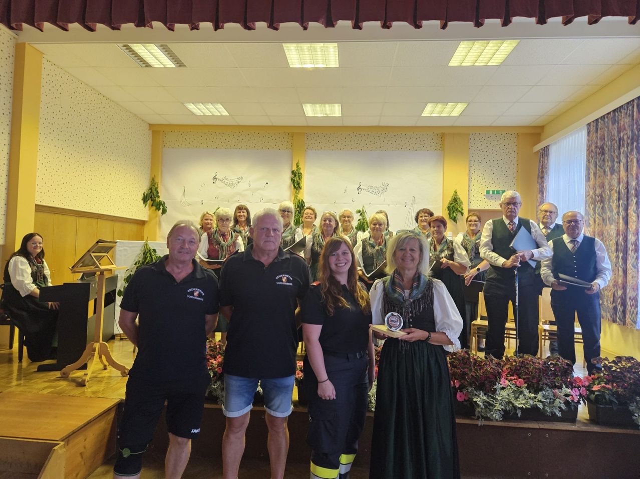 Eine Gruppe von Menschen steht in einem Raum mit Blumen und lächelt. Sie halten Auszeichnungen. Hinter ihnen steht eine Holzbühne. Der Raum hat eine weiße Wand mit Zeichnungen.