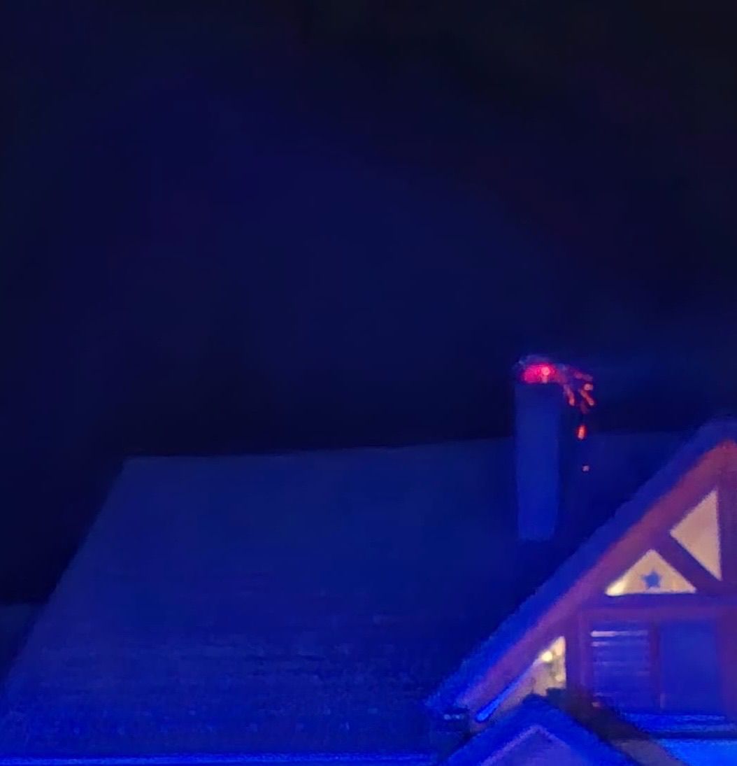 Ein Haus mit einem Schornstein und dreieckigen Fenstern ist bei Nacht von einem blauen Licht beleuchtet. Das Dach ist geneigt, und ein rotes Licht erscheint über dem Schornstein.