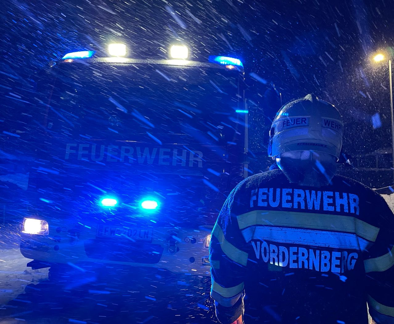 Ein Feuerwehrmann steht vor einem Feuerwehrauto in einer verschneiten Nacht. Die Scheinwerfer des Fahrzeugs beleuchten die Szene.