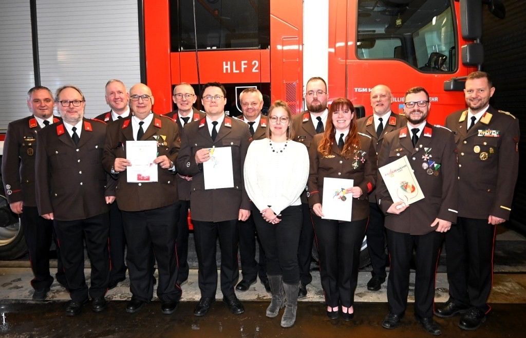 Eine Gruppe von Menschen in Uniformen, wahrscheinlich Feuerwehrleute, steht vor einem Feuerwehrfahrzeug mit der Bezeichnung HLF 2. Sie halten Urkunden in den Händen, und eine Frau in einem weißen Oberteil steht in der Mitte.