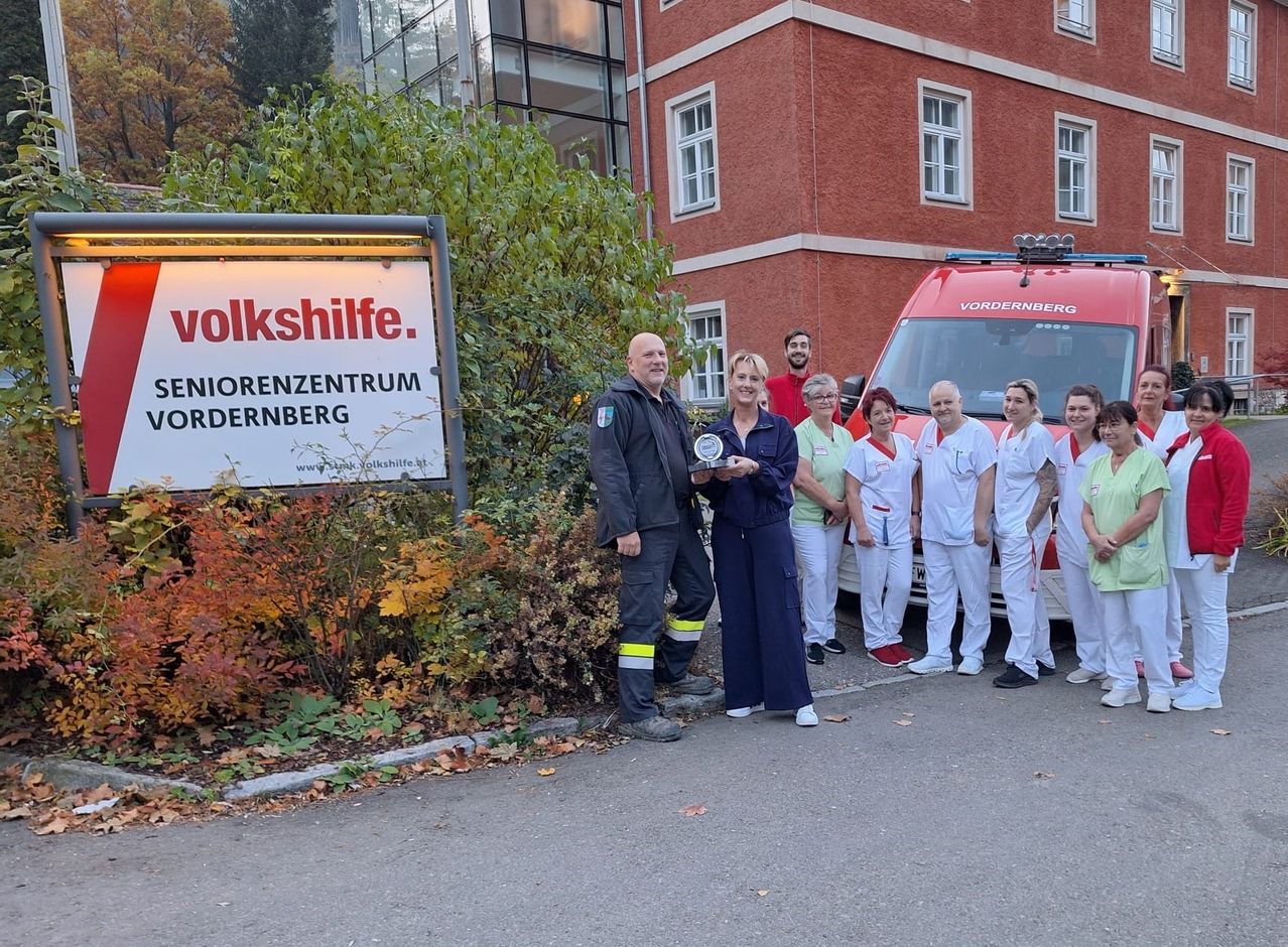 Eine Gruppe von Menschen in Uniform steht vor einem roten Krankenwagen, mit einem Schild für das Volkshilfe Seniorenzentrum Vordernberg im Hintergrund.