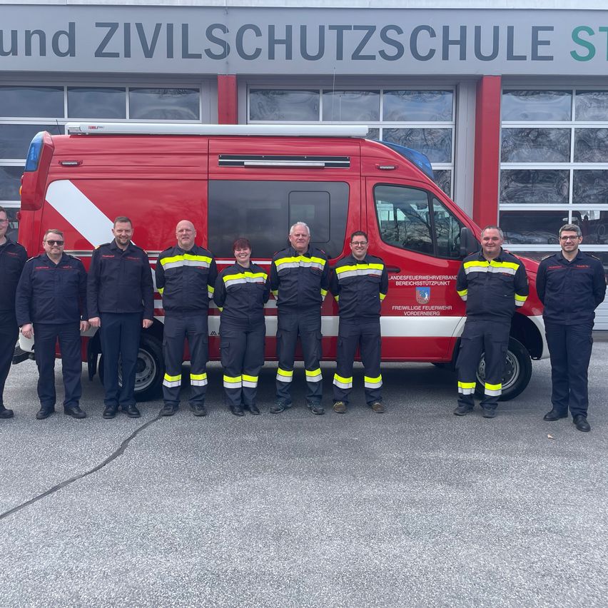 Eine Gruppe von Menschen in Uniformen steht vor einem roten Fahrzeug. Sie alle lächeln und posieren für ein Foto. Das Gebäude hinter ihnen hat ein Schild, auf dem 'Zivilschutzschule ST.' steht. Die Menschen tragen Uniformen, die schwarz und gelb sind.