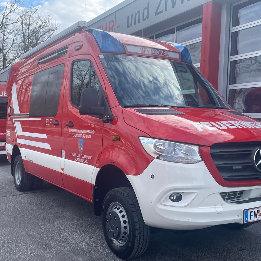 Ein roter und weißer Mercedes-Benz Feuerwehrwagen mit 'BFVLEOBEN' oben und 'FEUERWEHR' an der Seite steht vor einem Gebäude mit Glasfenstern.