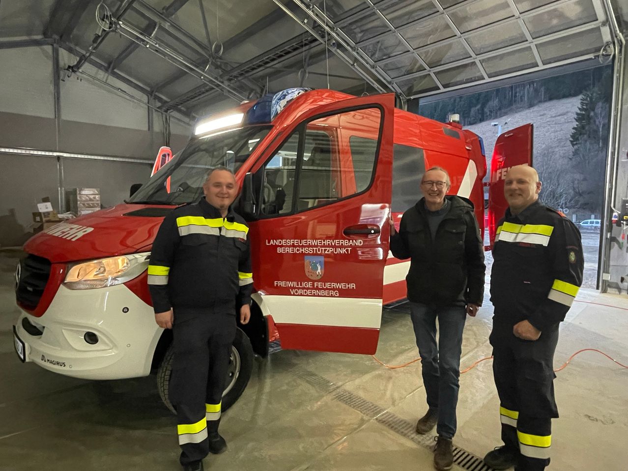 Drei Feuerwehrleute stehen neben einem roten Feuerwehrwagen in einer Garage und lächeln für ein Foto. Der Wagen trägt die Aufschrift 'Freiwillige Feuerwehr' und 'Vordereberg'.