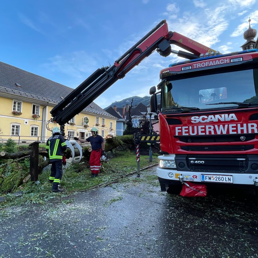 A red Scania fire truck is parked on the side of the road with its crane extended. Two firefighters are cutting down a fallen tree with chainsaws.
