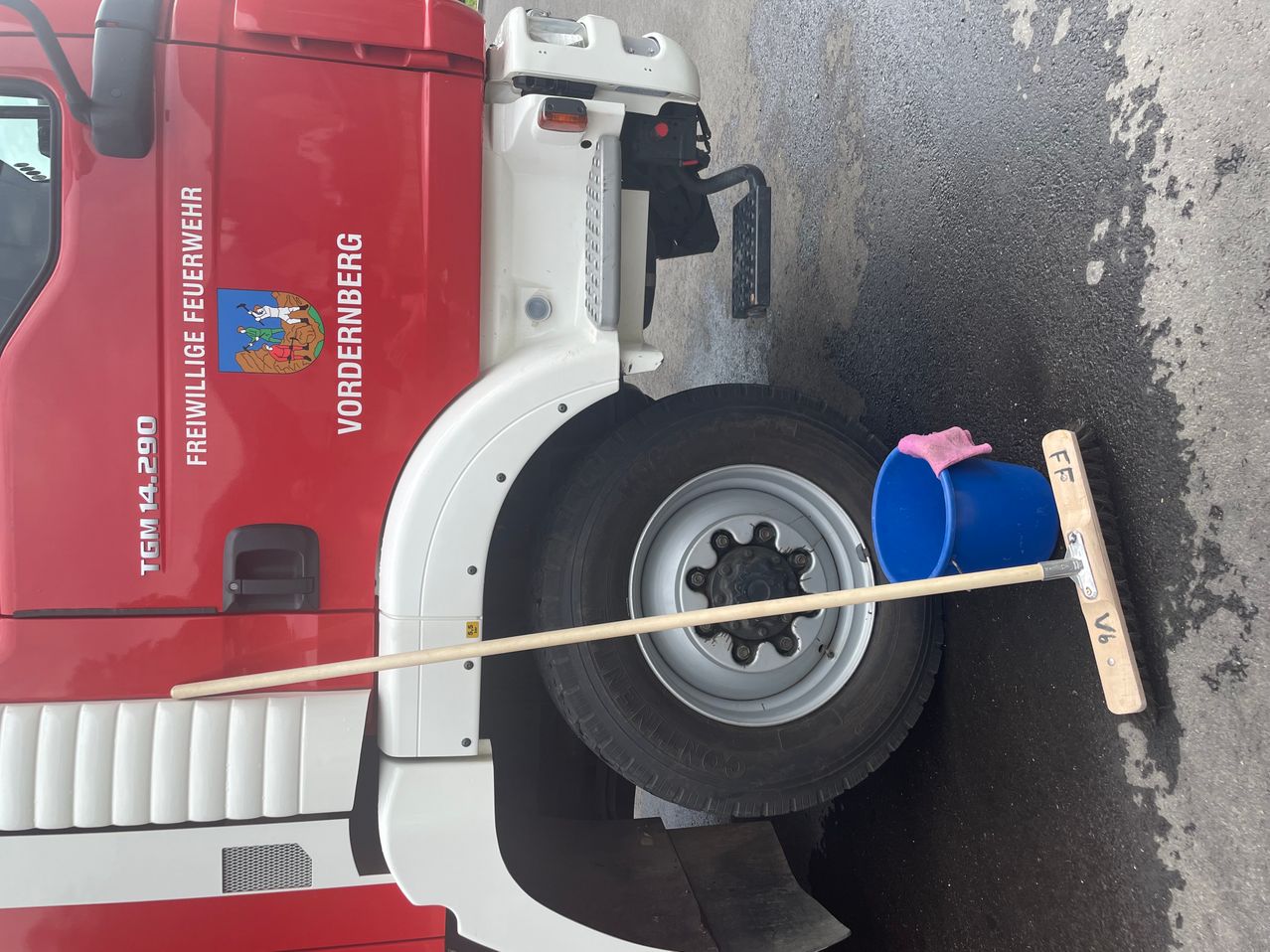 Ein roter Feuerwehrwagen mit dem Vordernberg-Emblem steht am Straßenrand. Ein blauer Eimer mit einem rosa Tuch steht in der Nähe des Rades.