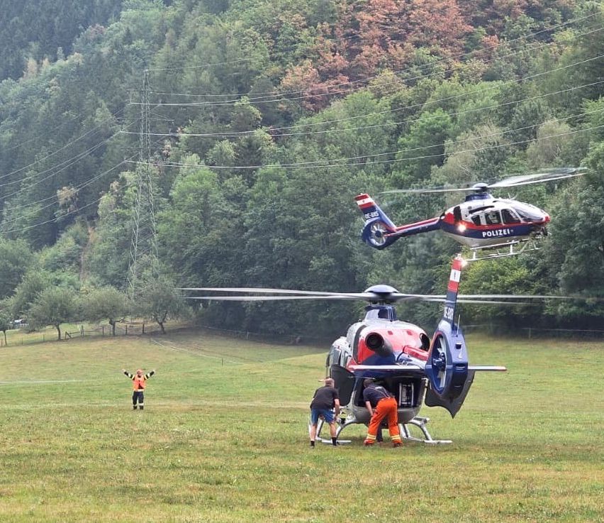 Zwei Polizeihubschrauber befinden sich auf einem Grasfeld, mit zwei Personen in orangefarbenen Anzügen in der Nähe. Ein Hubschrauber landet, während der andere über ihnen schwebt.