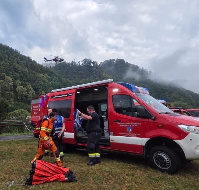 Rettungskräfte bereiten sich darauf vor, einer Person in Not zu helfen, während ein Hubschrauber über einem bergigen Gebiet fliegt.