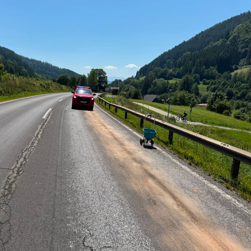 Ein rotes Auto fährt auf einer Bergstraße, während ein Radfahrer auf einem Seitenweg fährt. Die Straße wird von einem Schutzgeländer flankiert, und im Hintergrund befinden sich üppige grüne Hügel und ein klarer blauer Himmel.
