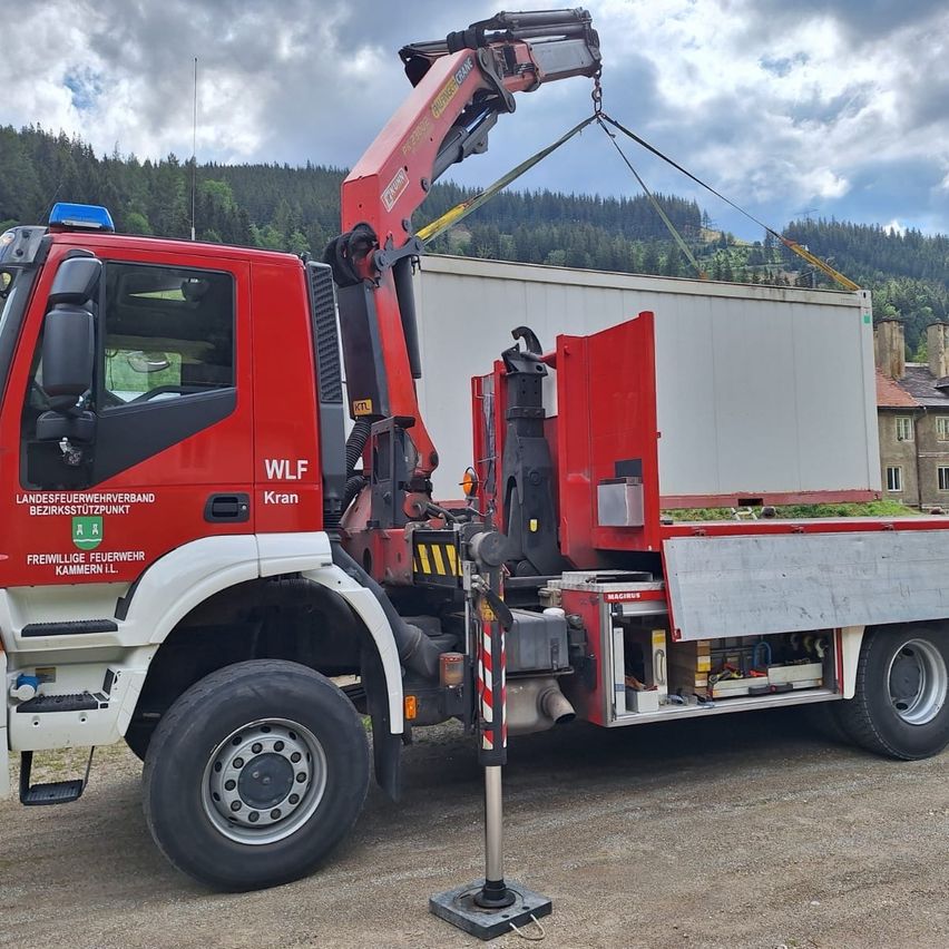 Ein roter Kranwagen mit der Aufschrift 'WLF Kran' hebt einen weißen Container. Der LKW ist auf einem unbefestigten Weg geparkt, mit einem Gebäude im Hintergrund.