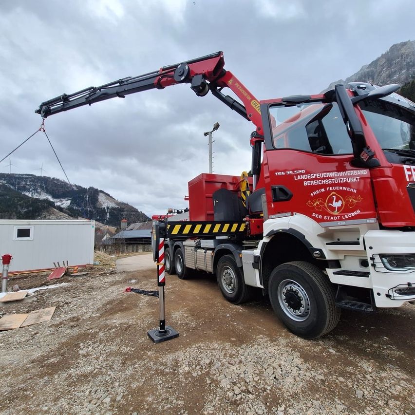 Ein roter und weißer LKW mit einem Kran ist auf einer Schotterstraße geparkt, möglicherweise für eine Baustelle. Der LKW ist mit den Texten 'Landesfeuerwehrverband' und 'Freiwillige Feuerwehr' beschriftet.