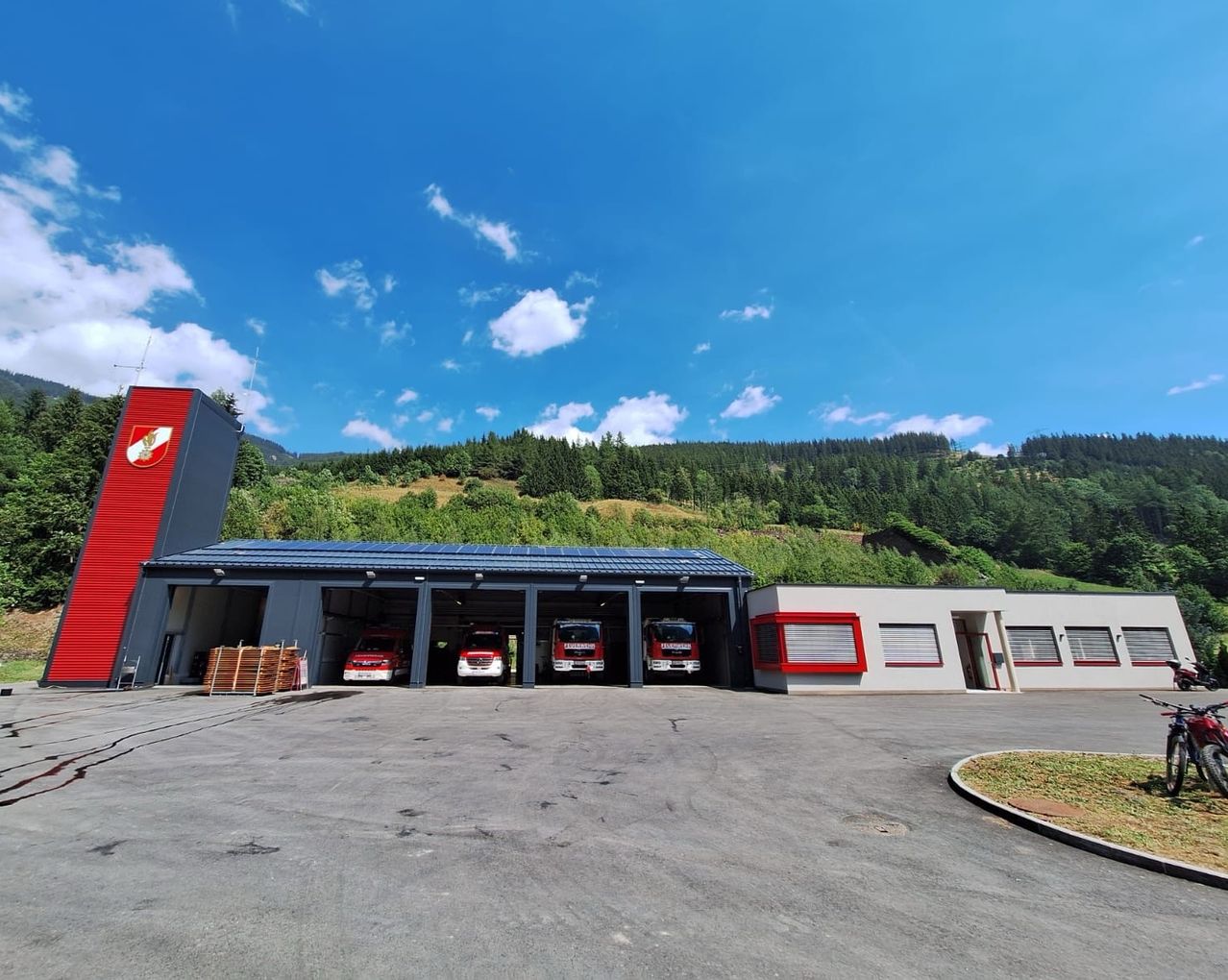 Eine Feuerwehrstation mit drei roten Feuerwehrfahrzeugen, die unter einem blauen Himmel mit einigen Wolken und einem Berg im Hintergrund geparkt sind.
