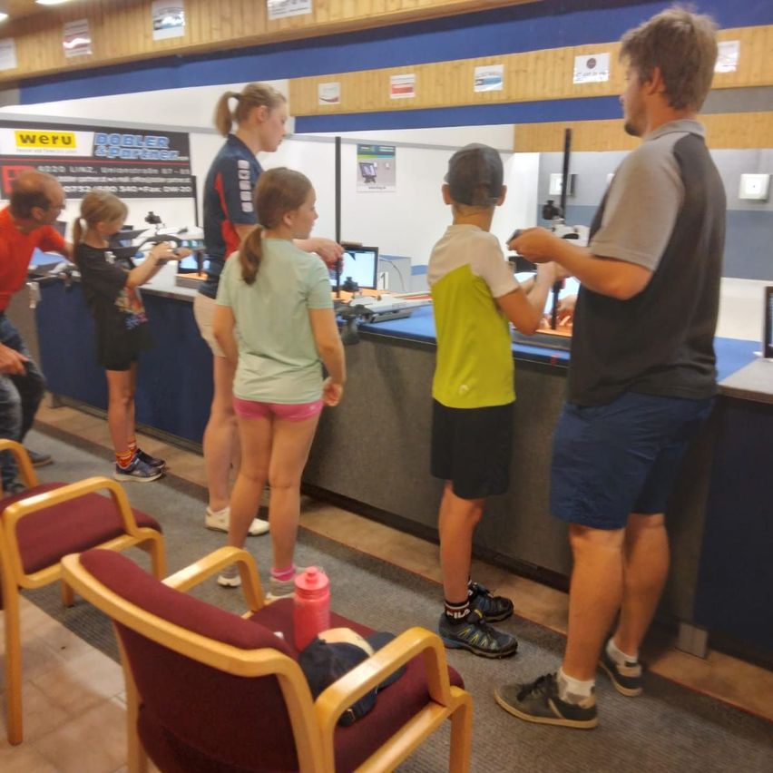 In einer Schießanlage zeigt ein Mann einer Gruppe von Kindern den Umgang mit Schusswaffen. Die Kinder stehen in einer Reihe, jede mit einer Waffe, und in der Nähe eines Tresens.