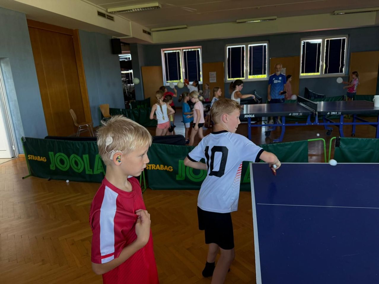 Zwei junge Jungen spielen Tischtennis in einer Turnhalle, während andere Kinder zuschauen. Der Junge in Rot schaut seinen Gegner an.