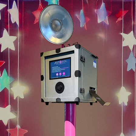 Ein Fotoautomat mit Bildschirm und Tasten steht auf einem rosa Stab vor einem Hintergrund aus bunten Sternen.