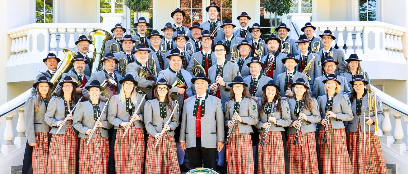 Eine Gruppe von Musikern in traditioneller Kleidung steht vor einem Gebäude. Sie halten verschiedene Instrumente, darunter Trompeten, Posaunen und Flöten. Vor ihnen steht ein Schlagzeug.