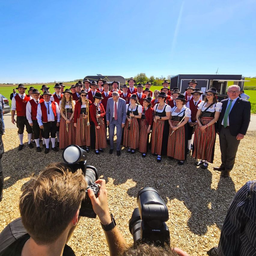 Eine Gruppe von Musikern in traditioneller bayerischer Kleidung, darunter auch einige Männer in Anzügen, sind auf einer Kiesfläche im Kreis versammelt, während ein Fotograf den Moment einfängt.