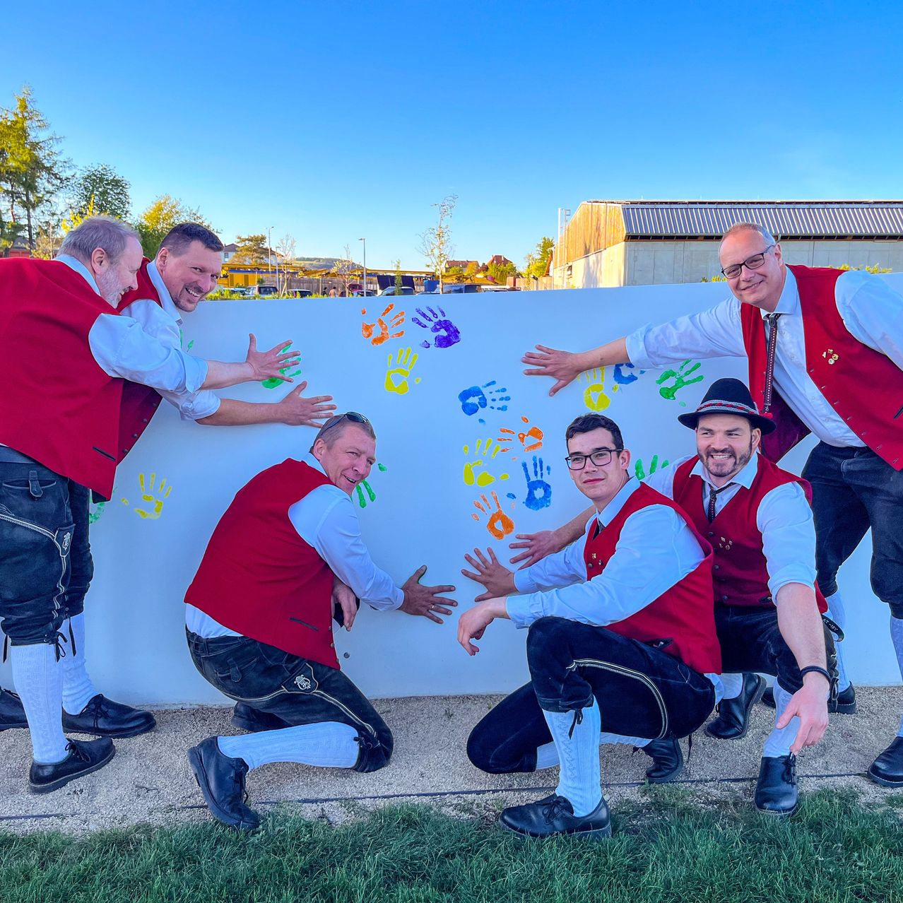 Fünf Männer in traditioneller bayerischer Kleidung posieren neben einer Wand mit bunten Handabdrücken und lächeln für ein Foto.