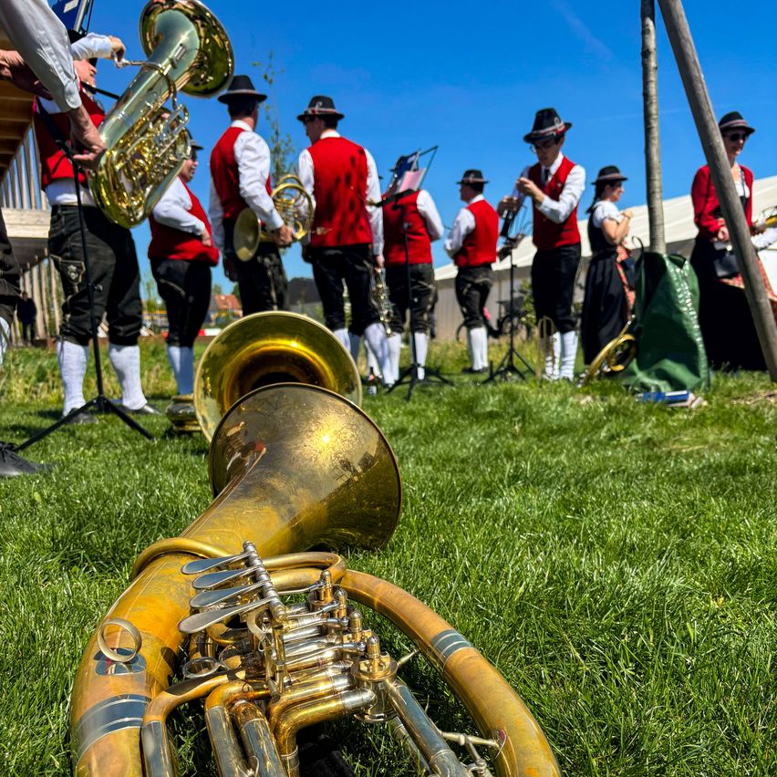 Eine Blaskapelle steht auf einem grünen Rasen, einige halten Instrumente. Die Musiker tragen rote Westen und schwarze Hosen. Der Tuba liegt auf dem Gras.