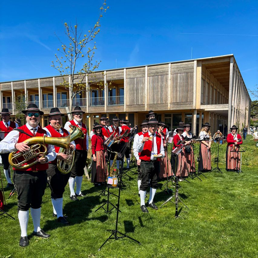 Eine Band in traditioneller Kleidung spielt auf einem Rasenfeld vor einem Holzgebäude an einem blauen Himmel.