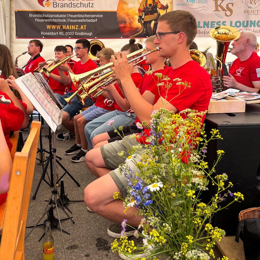 Eine Gruppe von Musikern, die rote T-Shirts tragen, sitzen auf Stühlen und spielen Trompeten und Posaunen. Sie befinden sich in einer Außenumgebung mit einem Banner im Hintergrund.