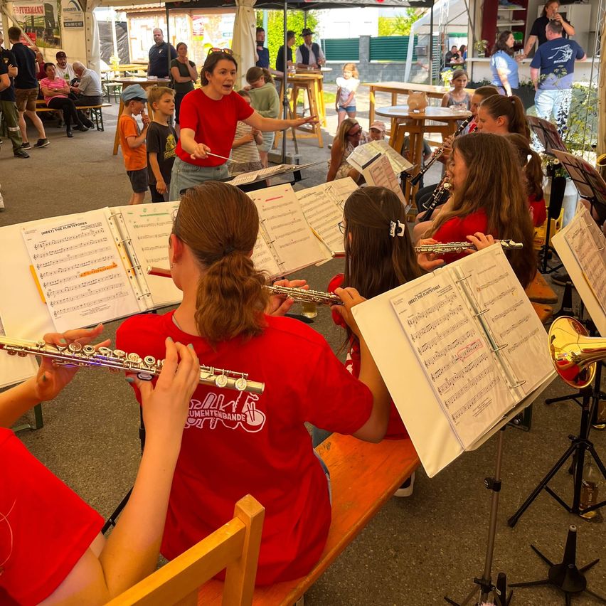 Eine Gruppe junger Musiker spielt ihre Instrumente unter der Anleitung einer Lehrerin, die in einem Zelt sitzt. Sie halten ihre Notenblätter und scheinen sich auf ihre Übung zu konzentrieren.