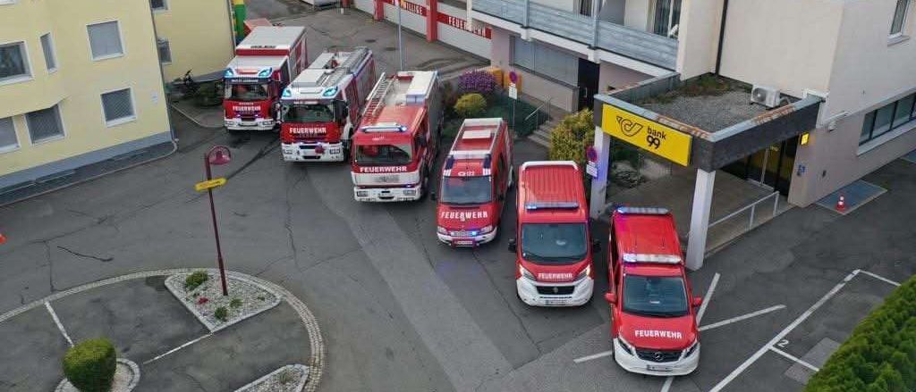 Eine Luftaufnahme einer Feuerwehrstation mit mehreren roten Feuerwehrfahrzeugen, die vor dem Gebäude geparkt sind. Eine Fahne ist auf einem Mast gehisst.