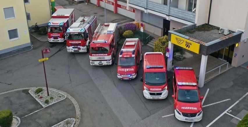 Eine Luftaufnahme einer Feuerwehrstation mit mehreren roten Feuerwehrfahrzeugen, die vor dem Gebäude geparkt sind. Eine Fahne ist auf einem Mast gehisst.