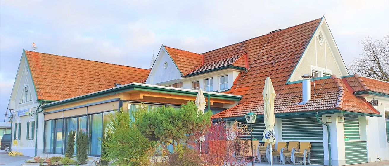 Ein modernes Gebäude mit roten Dächern und grüner Einfassung hat Glasfenster und einen Eingang mit einem Baldachin. Draußen stehen gelbe Stühle und Tische und ein geschlossener Schirm.