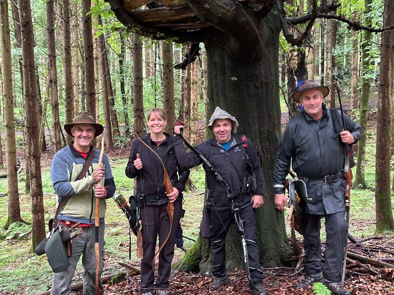 Vier Personen stehen in einem Wald, zwei Männer und zwei Frauen, alle lächeln. Sie tragen Outdoor-Ausrüstung und halten Bögen und Pfeile. Hinter ihnen ein Baumstamm mit einer Holzplattform obenauf.