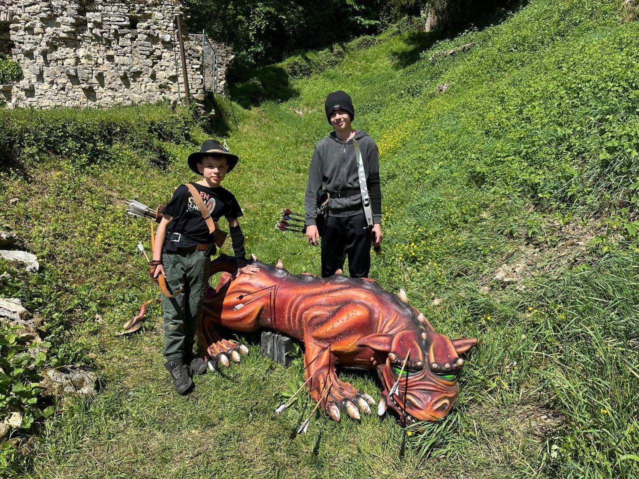 Zwei Kinder stehen neben einer drackenförmigen Requisite mit Pfeilen darin. Der mit dem Hut hält einen Bogen. Hinter ihnen ist eine Steinmauer.