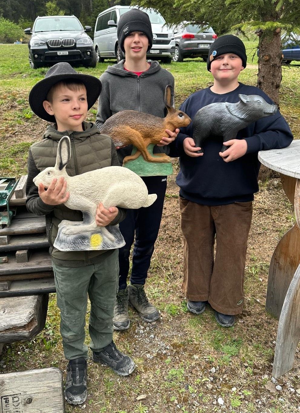 Three boys stand in a grassy area. The boy in the middle holds a brown rabbit statue, while the boy on the left holds a white rabbit statue and the boy on the right holds a gray wild boar statue.
