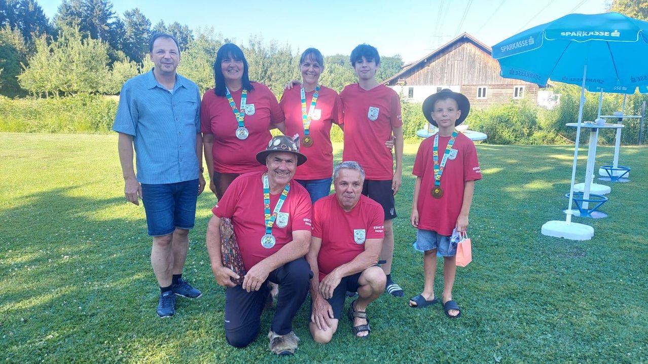 A group of people wearing red shirts and medals pose for a photo on a grassy field with a barn in the background.
