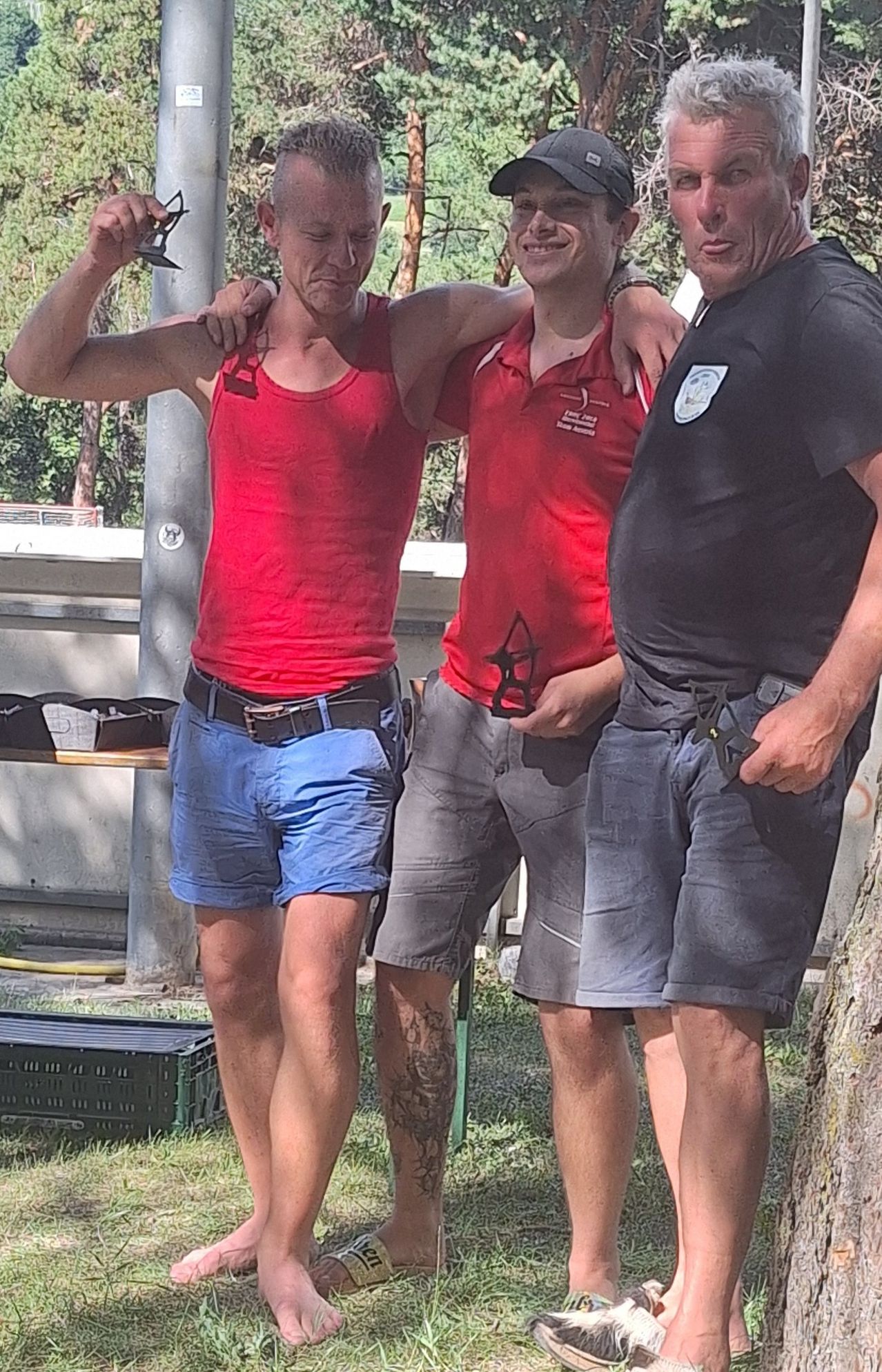 Three men stand together, one wearing a red shirt, another in a black shirt, and the third in a red shirt with a logo on the left chest. They are smiling and appear to be posing for a photo.