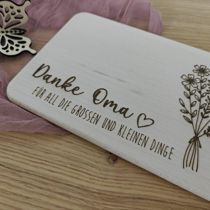 Eine Holztafel mit Blumen und den Worten 'Danke Oma' ist auf einer Holzoberfläche ausgestellt. Die Nachricht 'für all die großen und kleinen Dinge' ist in kursiver Schrift geschrieben. Ein Schmetterling ist links.