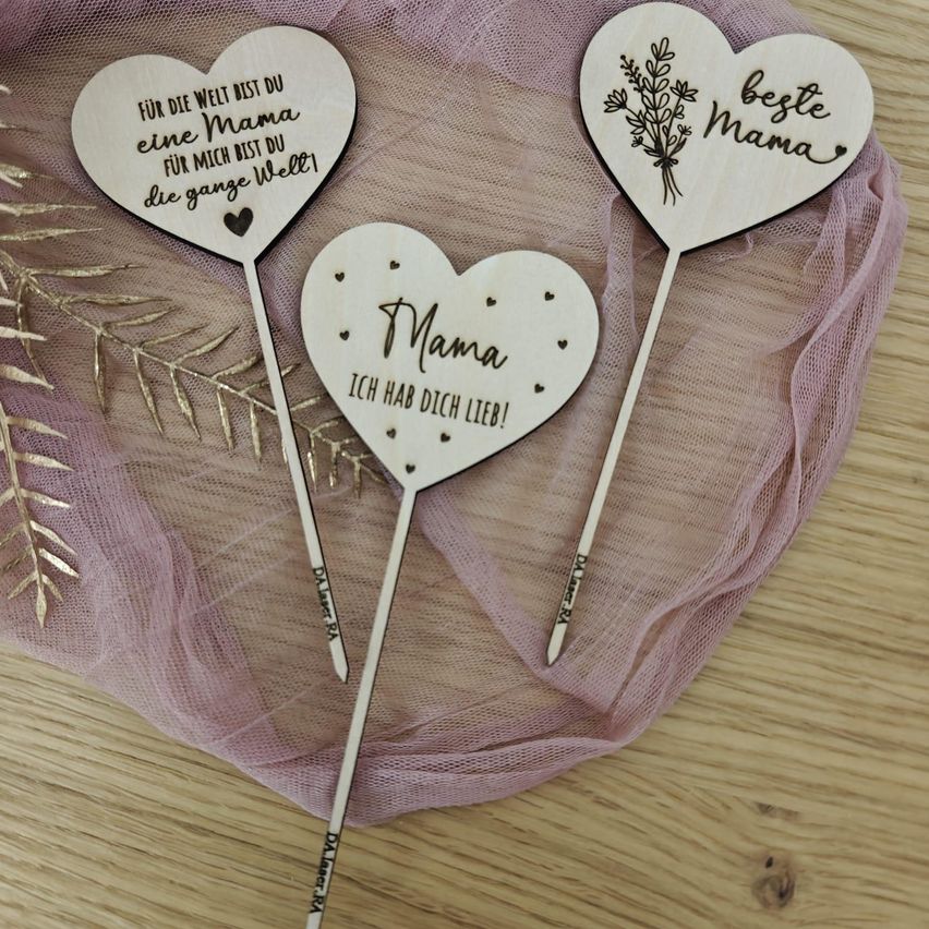 Drei herzförmige Holzstäbe mit dem Text 'Für die Welt bist du die beste Mama, ich liebe dich so sehr' auf einem rosa Tuch.