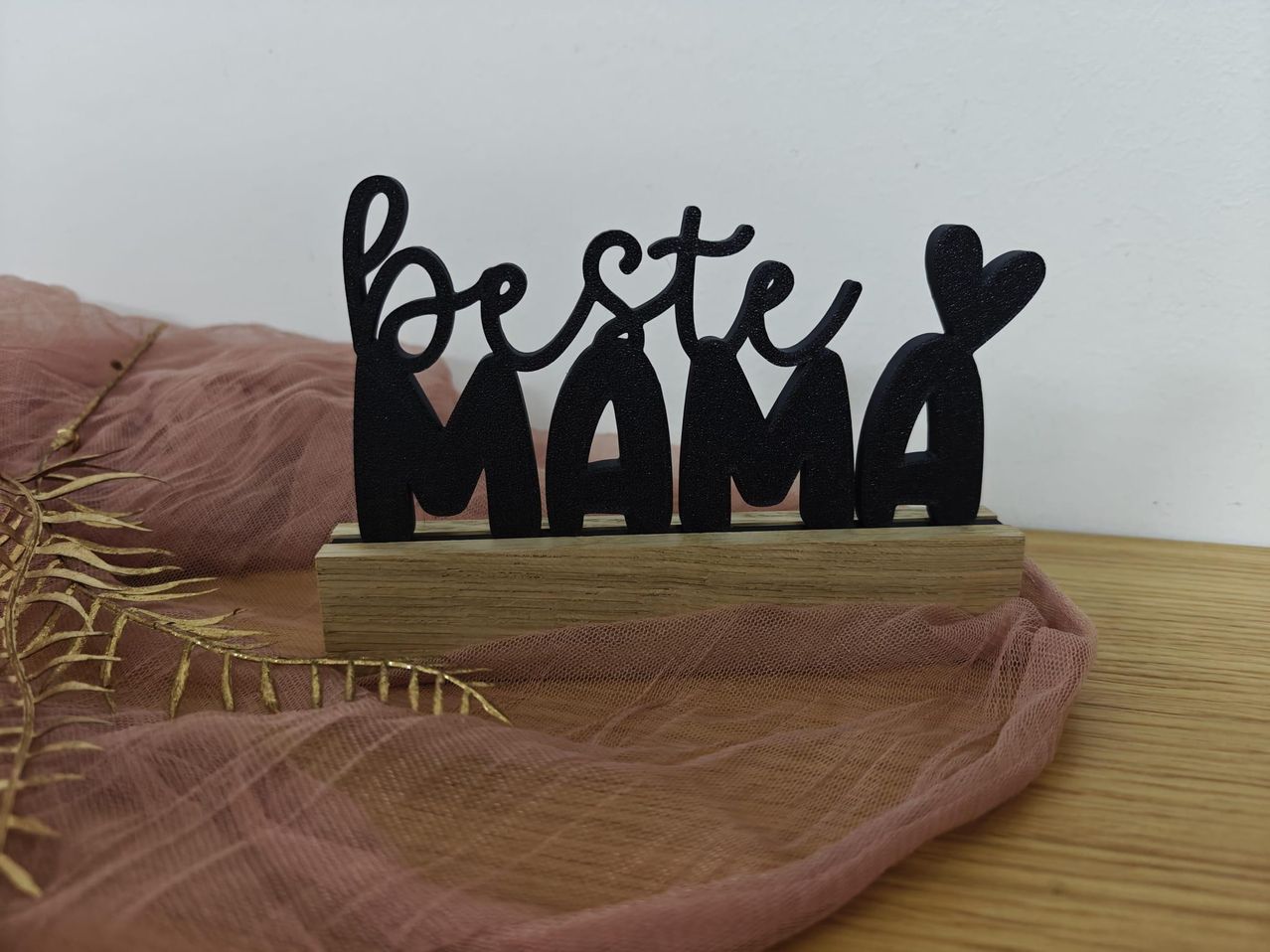 Ein Holzständer zeigt ein schwarzes Schild mit den Worten 'Beste Mama' in kursiver Schrift. Zwei Herzformen befinden sich auf beiden Seiten des Textes. Ein rosa Tuch bedeckt den unteren Teil.