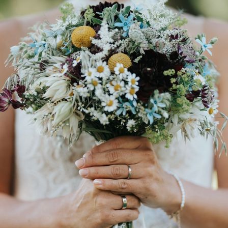 Eine Braut hält einen Hochzeitsstrauß mit weißen, blauen und gelben Blumen und grünen Blättern.